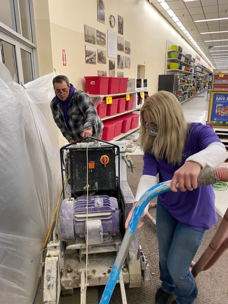 Two people, wearing casual clothes, operate a floor grinding machine inside a store with shelves and red bins behind them.