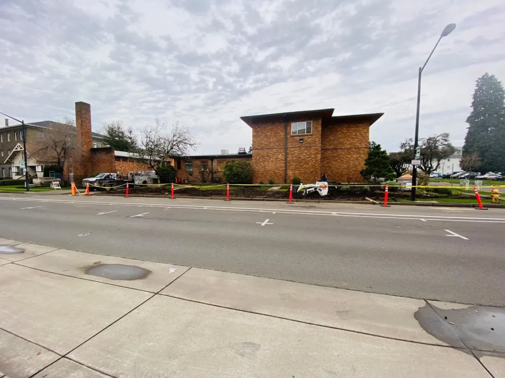 Street view of a brick building under construction with orange safety cones and a fenced-off area in front.