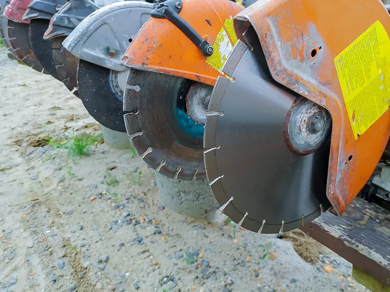 Close-up of a circular saw blade attached to an orange saw, placed on a sandy surface.