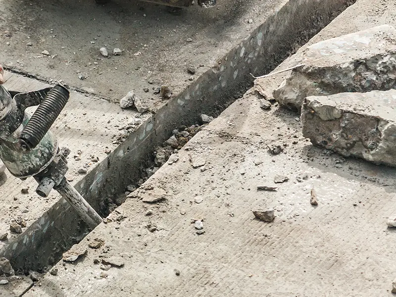 A worker uses a jackhammer to break concrete, creating a groove between slabs, with scattered debris around.