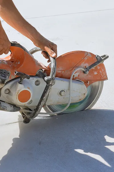 Close-up of a person operating an orange concrete saw, cutting through a white concrete surface.